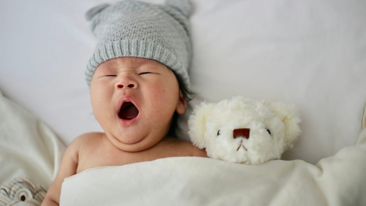 Bebé de 6 meses bostezando en la cama junto a un osito de peluche